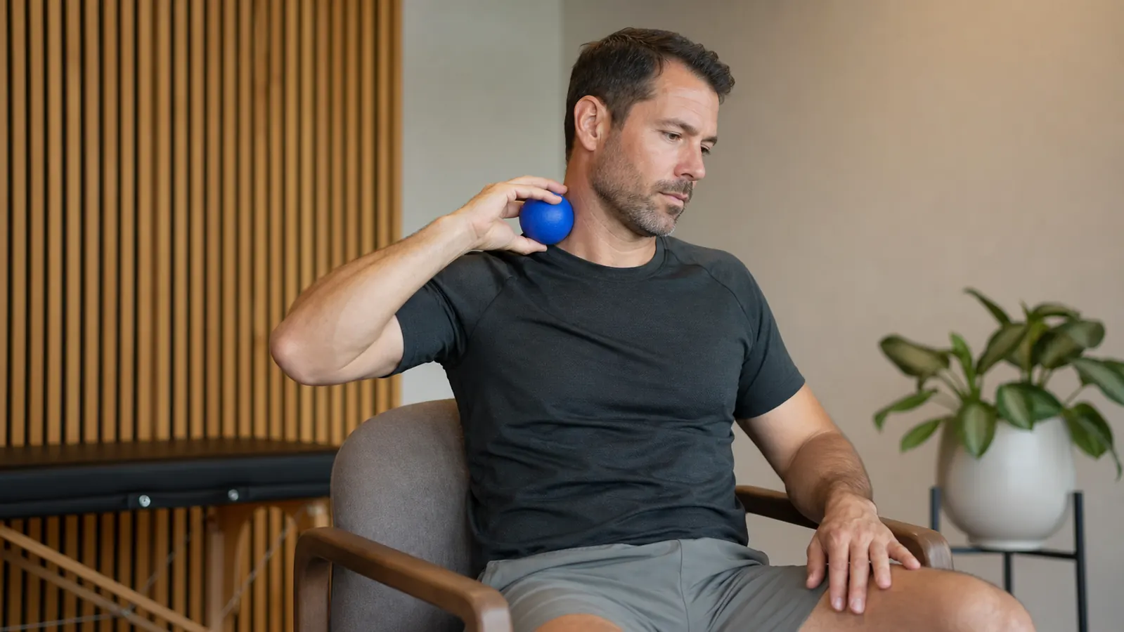 Man pressing a lacrosse ball into the base of his neck against a gym wall, eyes closed, focused